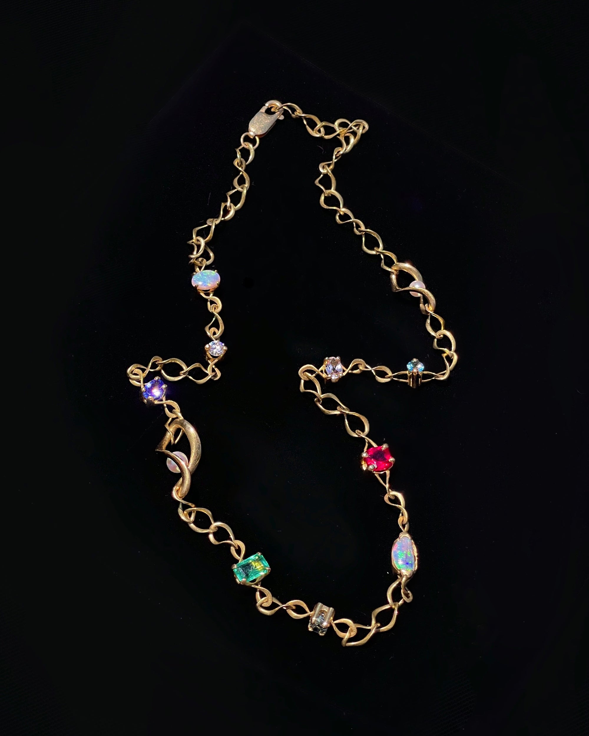 A gold chain necklace featuring multiple colorful gemstone accents in purple, green, blue, and red, displayed against a solid black background.